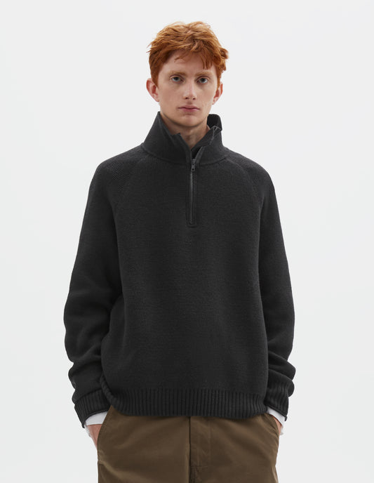 Cropped body image, front view of model wearing black wool cotton zip up polo jumper with contrast zip on collar, raglan sleeve and contrast rib on cuffs and hem. Styled with white shirt and khaki green trousers.