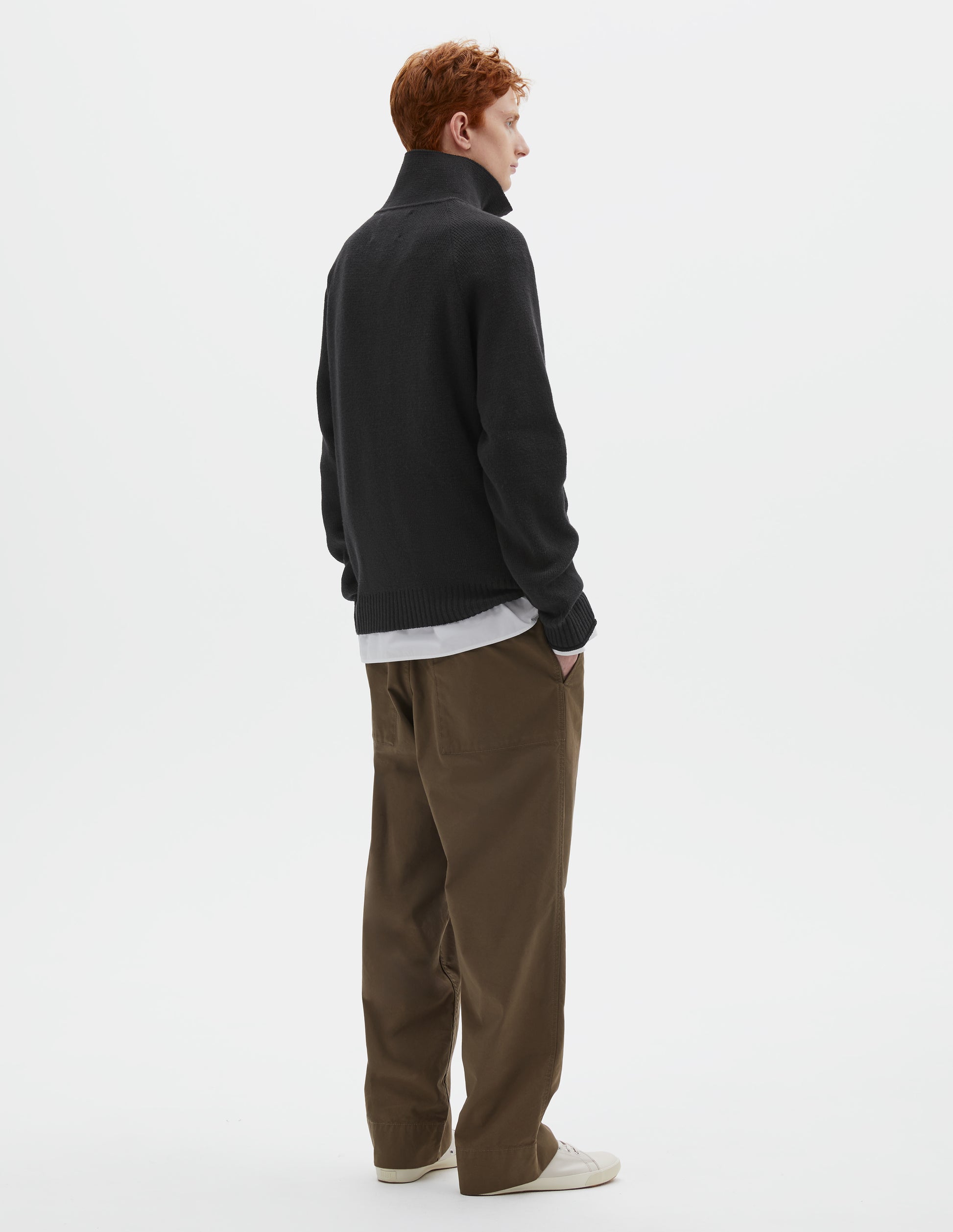 Full body image, back view of model wearing black wool cotton zip up polo jumper, with raglan sleeve and contrast rib on cuffs and hem. Styled with white shirt, khaki green trousers and beige plimsolls.