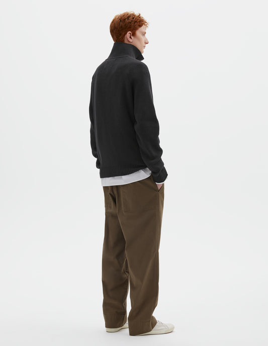 Full body image, back view of model wearing black wool cotton zip up polo jumper, with raglan sleeve and contrast rib on cuffs and hem. Styled with white shirt, khaki green trousers and beige plimsolls.