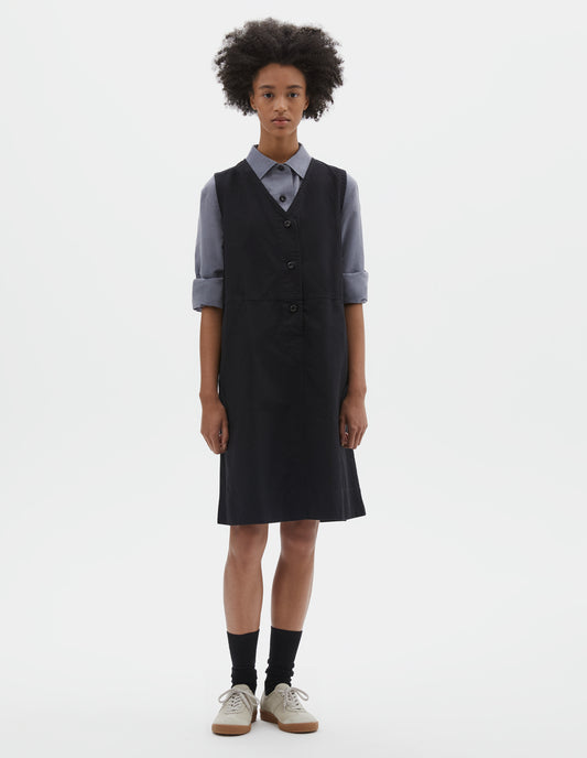 Full body image, front view of model wearing black sleeveless dress with v neck detailing, slant side pockets and corozo brace button closure. Styled over blue cotton shirt, with black socks and beige suede trainers.