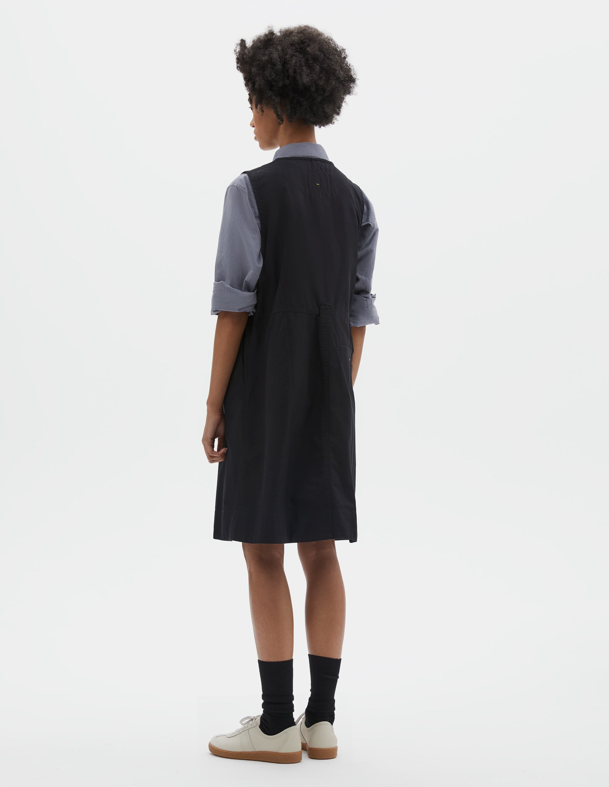 Full body image, back view of model wearing black sleeveless dress with singular belt loop and back button through welt pocket. Styled over blue shirt, with black socks and beige suede trainers.