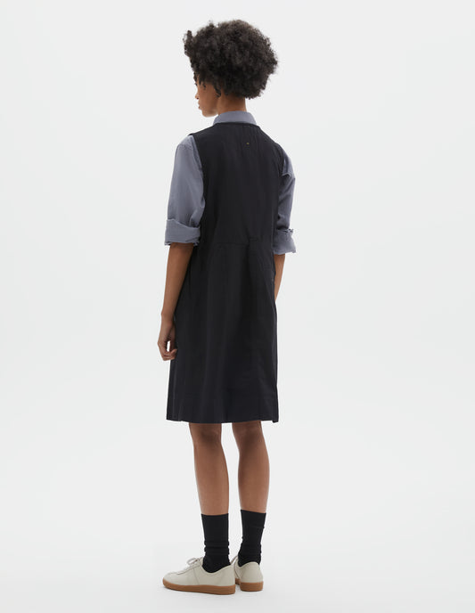 Full body image, back view of model wearing black sleeveless dress with singular belt loop and back button through welt pocket. Styled over blue shirt, with black socks and beige suede trainers.