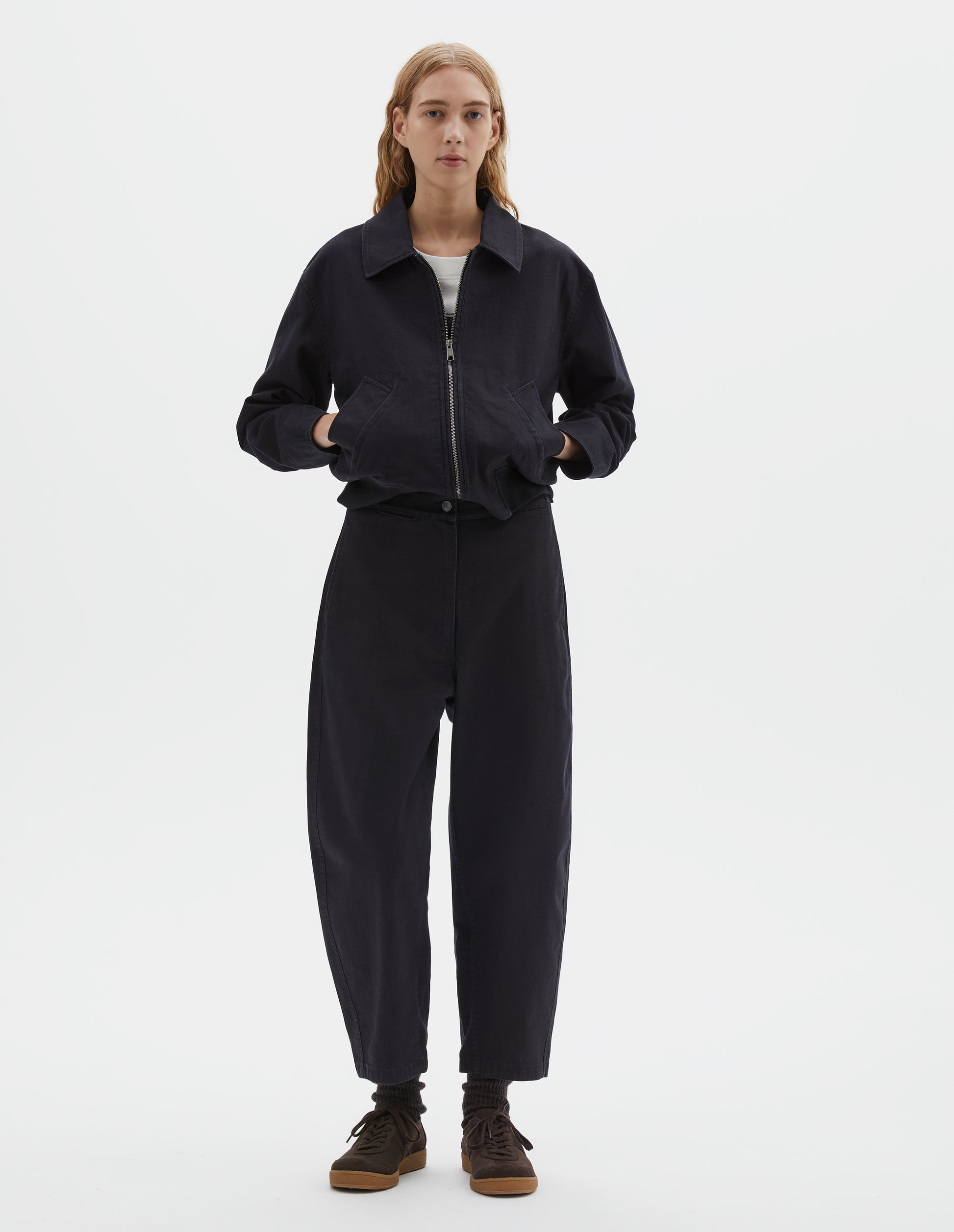 Full body image, front view of model wearing indigo blue barrel leg trouser, with slant side pockets and silver metal closure. Styled with indigo blue jacket, white t-shirt, black socks and brown suede trainers. 