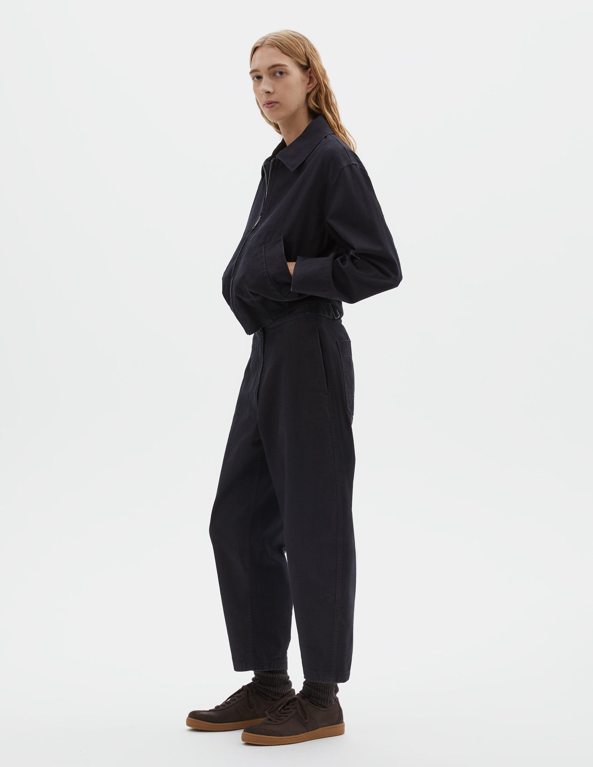 Full body image, front view of model wearing cropped boxy dark blue jacket with zip fastening, deep slanted welt pockets with reinforced stitch detailing and rib elasticated waistband. Styled with matching trousers and dark brown socks with dark brown nubuck trainers.