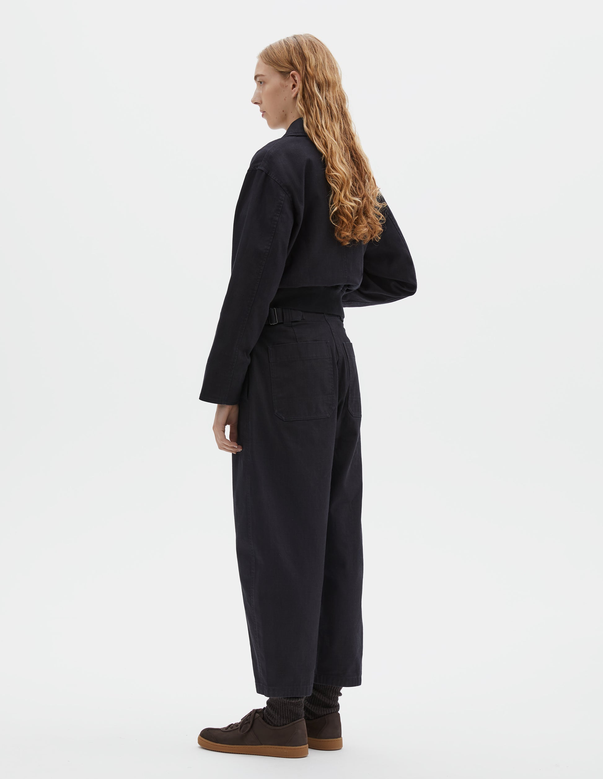 Full body image, back view of model wearing indigo blue barrel leg trouser, with two patch pockets and silver metal waist tab adjuster. Styled with indigo blue jacket, white t-shirt, black socks and brown suede trainers.