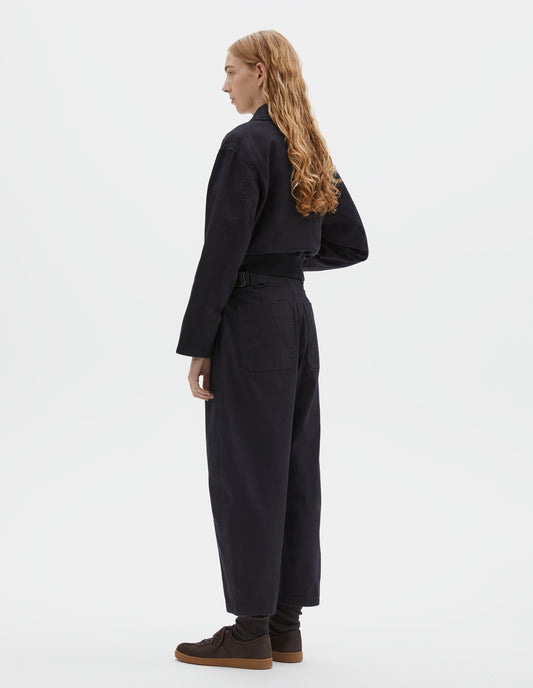 Full body image, back view of model wearing indigo blue barrel leg trouser, with two patch pockets and silver metal waist tab adjuster. Styled with indigo blue jacket, white t-shirt, black socks and brown suede trainers.