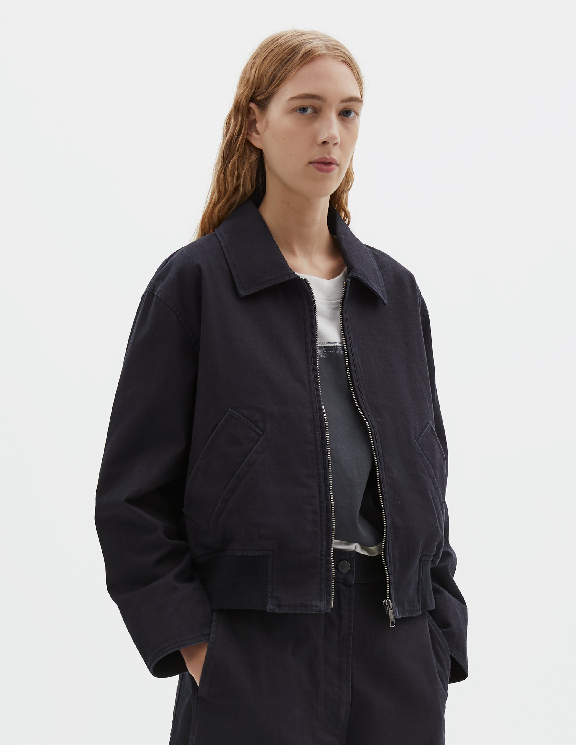 Cropped body image, front view of model wearing cropped boxy dark blue jacket with zip fastening, deep slanted welt pockets with reinforced stitch detailing and rib elasticated waistband. Styled over white t-shirt with black print, and with matching trousers.