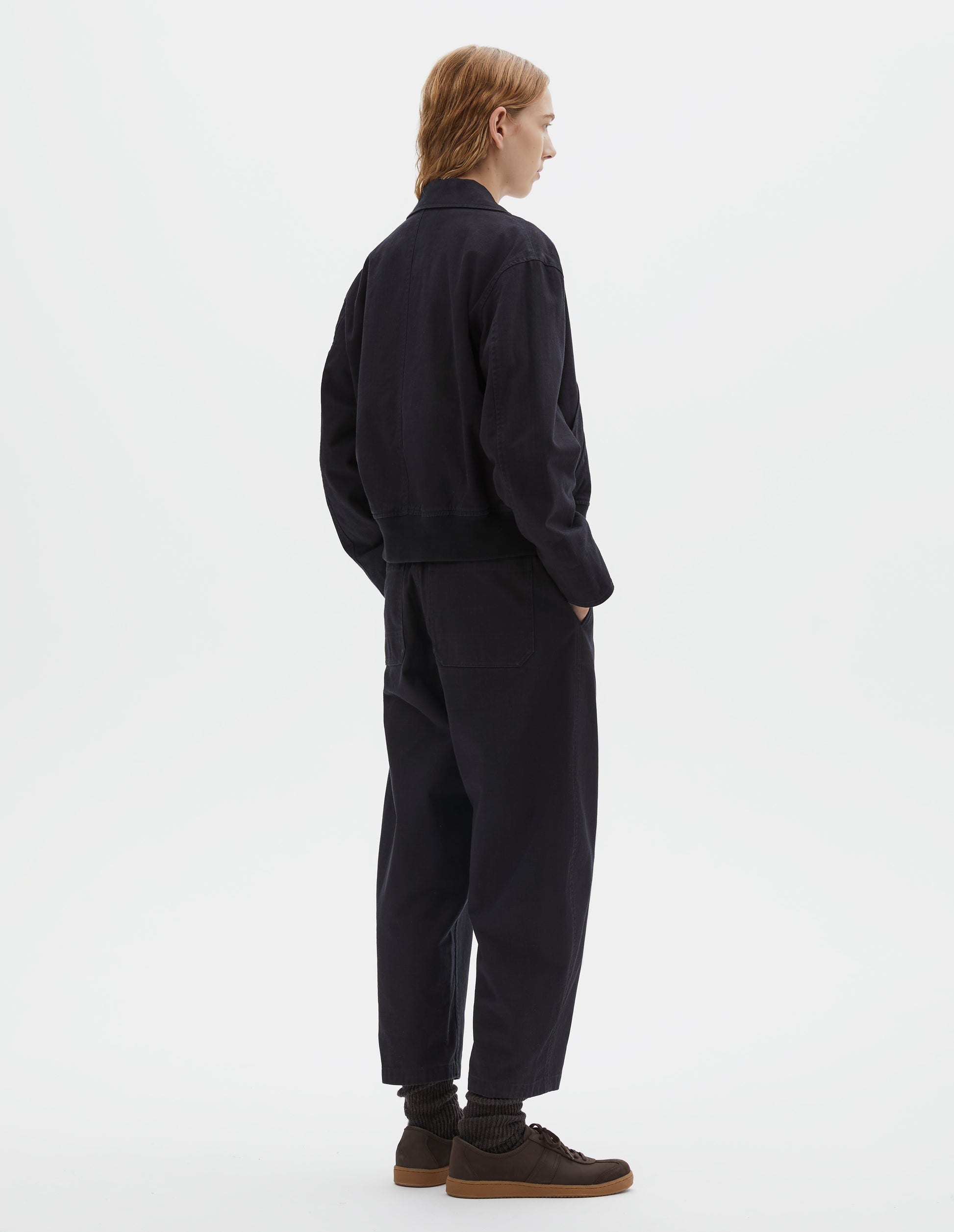 Full body image, back view of model wearing cropped boxy dark blue jacket with rib elasticated waistband. Styled with matching trousers and dark brown socks with dark brown nubuck trainers.