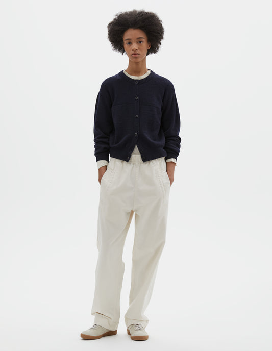 Full body image, front view of model wearing dark navy blue wool textured cardigan, with contrast panel across chest, rib detailing on neck, cuffs and hem and corozo button closure. Styled over off white long sleeve t shirt, off white joggers and beige suede trainers.