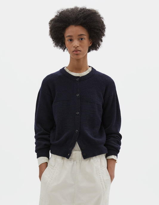 Cropped body image, front view of model wearing dark navy blue wool textured cardigan, with contrast panel across chest, rib detailing on neck, cuffs and hem and corozo button closure. Styled over off white long sleeve t shirt and off white joggers.