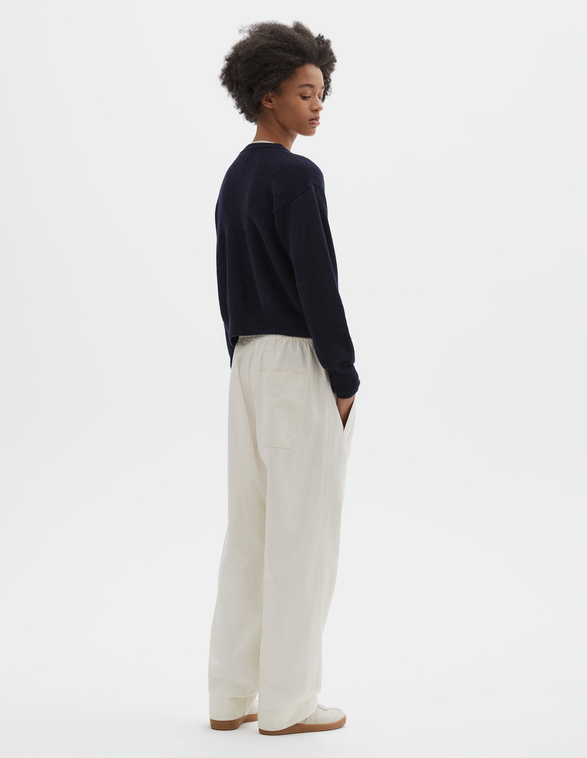 Full body image, back view of model wearing dark navy blue wool textured cardigan, with contrast panel across upper back and rib detailing on neck, cuffs and hem. Styled with off white joggers and beige suede trainers.