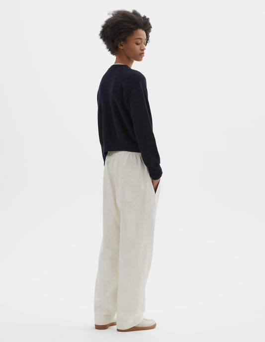 Full body image, back view of model wearing dark navy blue wool textured cardigan, with contrast panel across upper back and rib detailing on neck, cuffs and hem. Styled with off white joggers and beige suede trainers.
