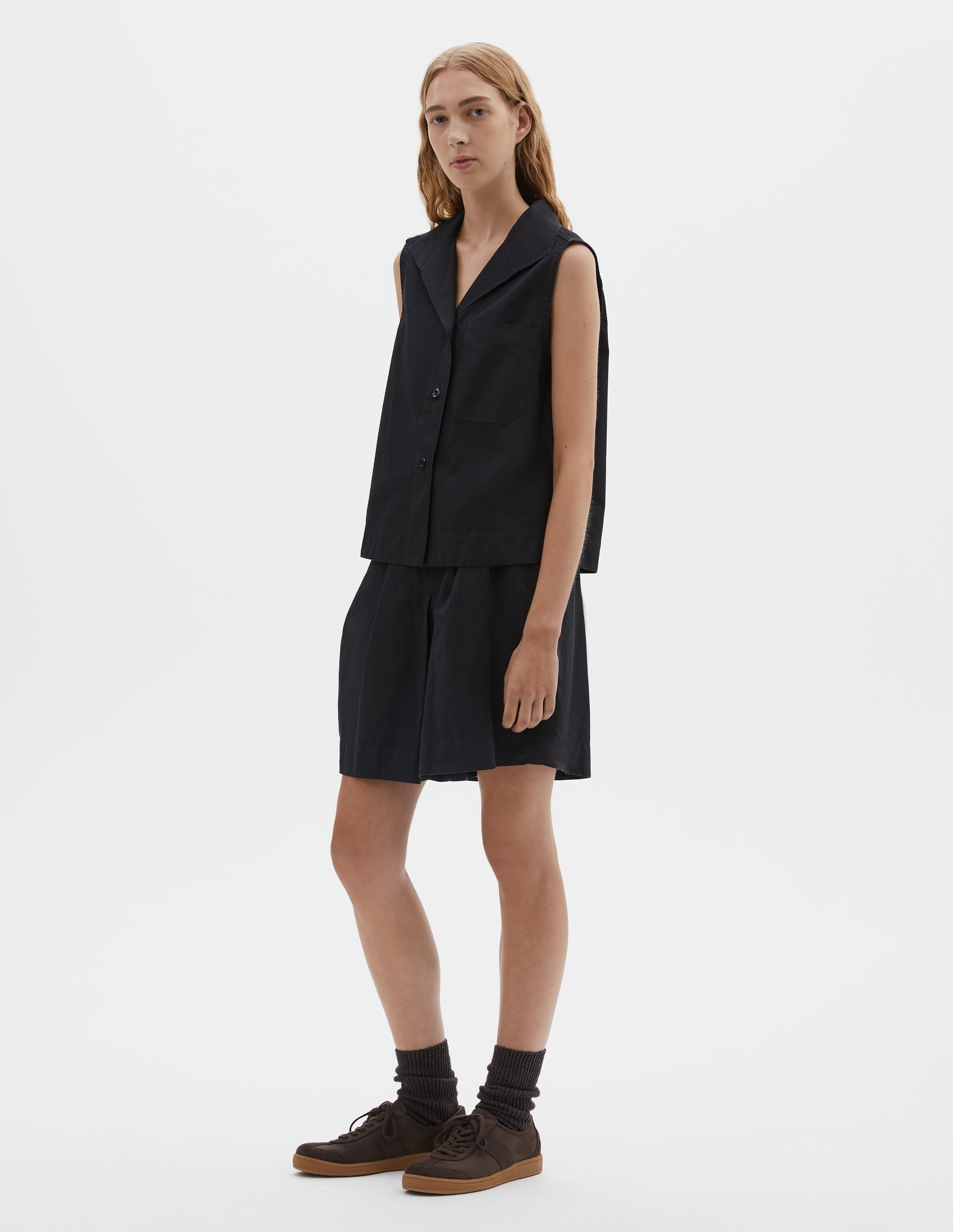 Full body image, front view of model wearing black denim sleeveless shirt with one-piece open spread collar, wide stitch-through placket, corozo brace button closure and single chest pocket. Styled with black shorts, dark brown socks and dark brown nubuck trainers.