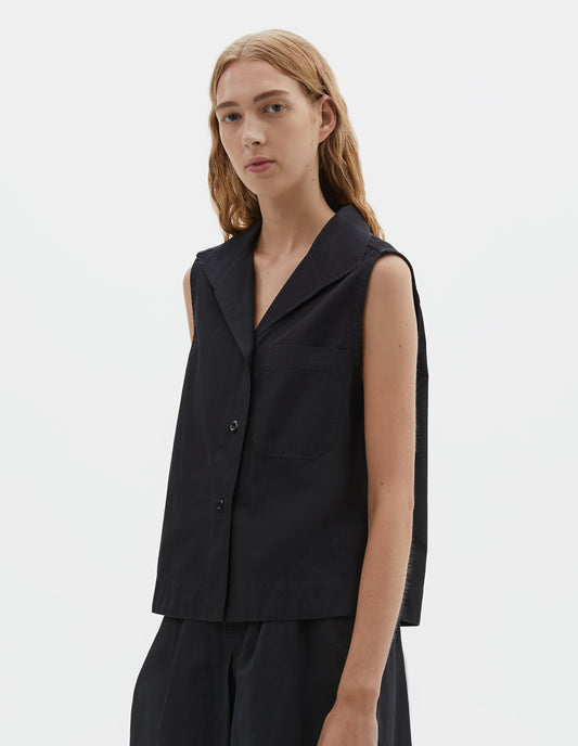 Cropped body image, front view of model wearing black denim sleeveless shirt with one-piece open spread collar, wide stitch-through placket, corozo brace button closure and single chest pocket. Styled with black shorts. 