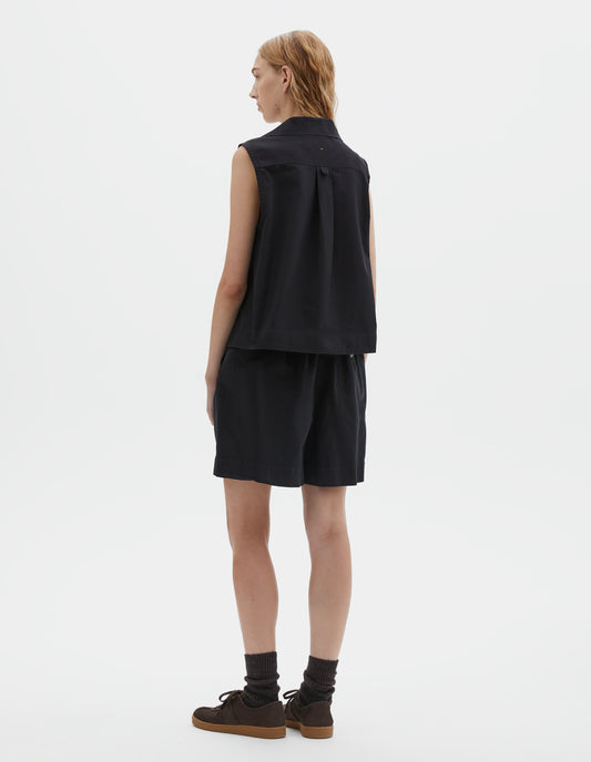 Full body image, back view of model wearing black denim sleeveless shirt with box pleat and hanger loop detail on back yoke. Styled with black shorts, dark brown socks and dark brown nubuck trainers.