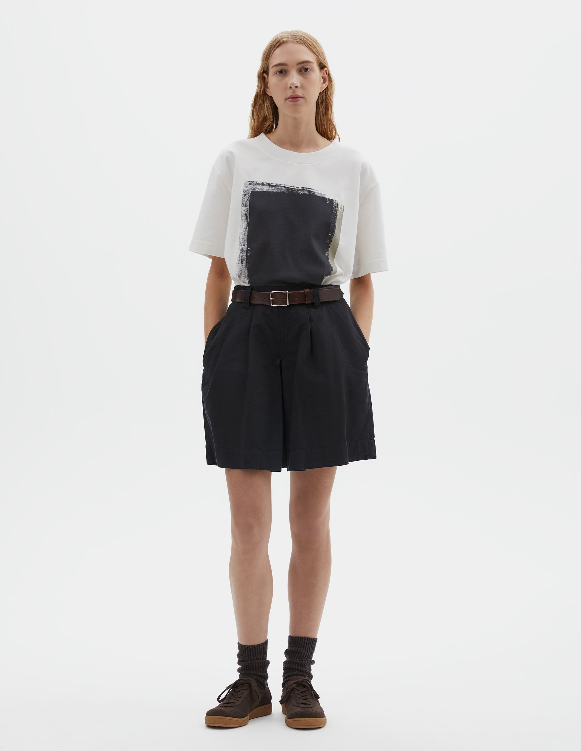 Full body image, front view of model wearing black cotton linen shorts with pleated skirt front panel, semi-elasticated waistband, side pockets, and belt loops. Styled with white t-shirt with black print, brown leather belt, brown socks and brown suede trainers with gum sole.