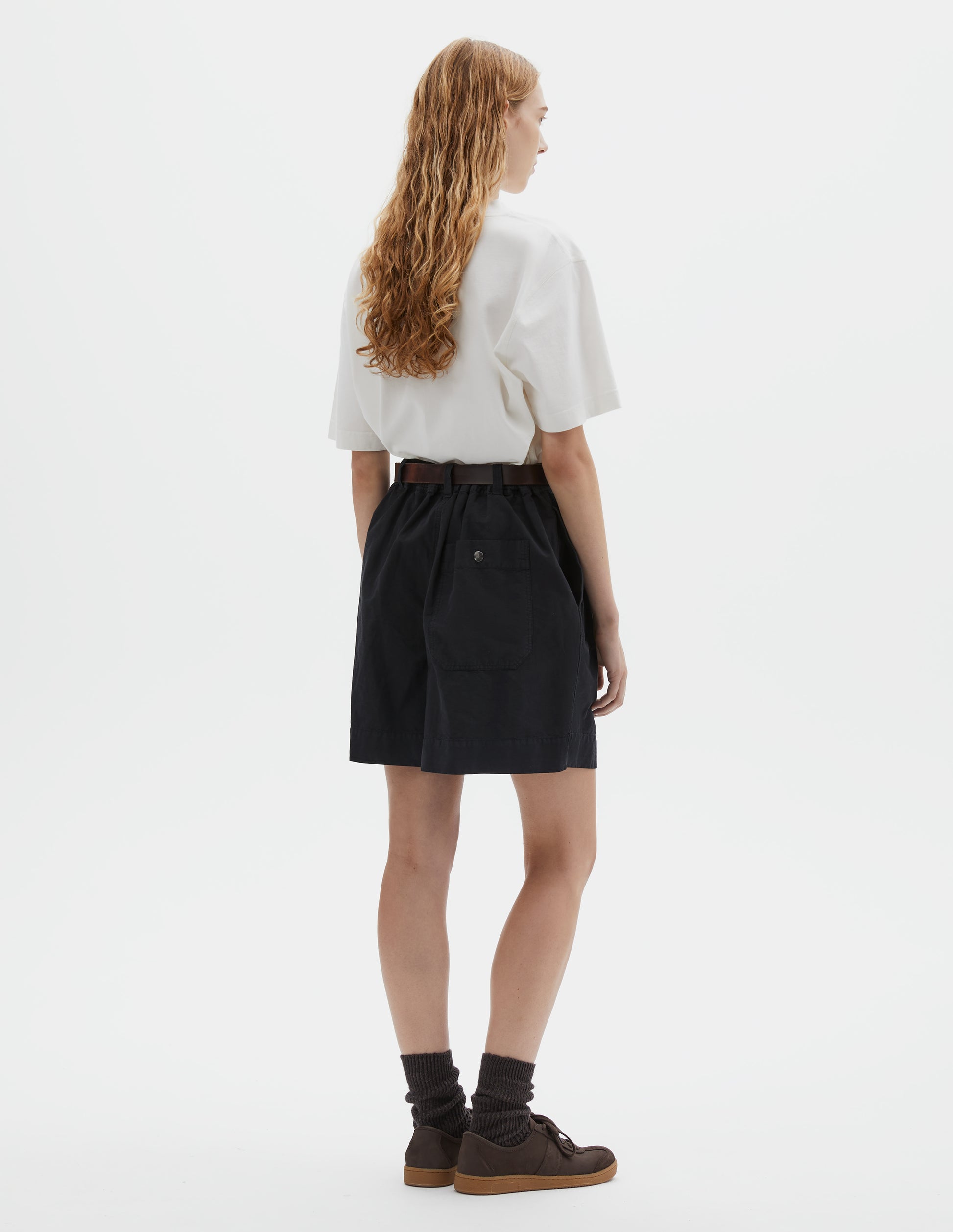 Full body image, back view of model wearing black shorts with semi-elasticated waistband, press stud fastening on back patch pocket and belt loops with one D-ring detail. Styled with white t-shirt, brown leather belt, brown socks and brown suede shoes with gum sole. 