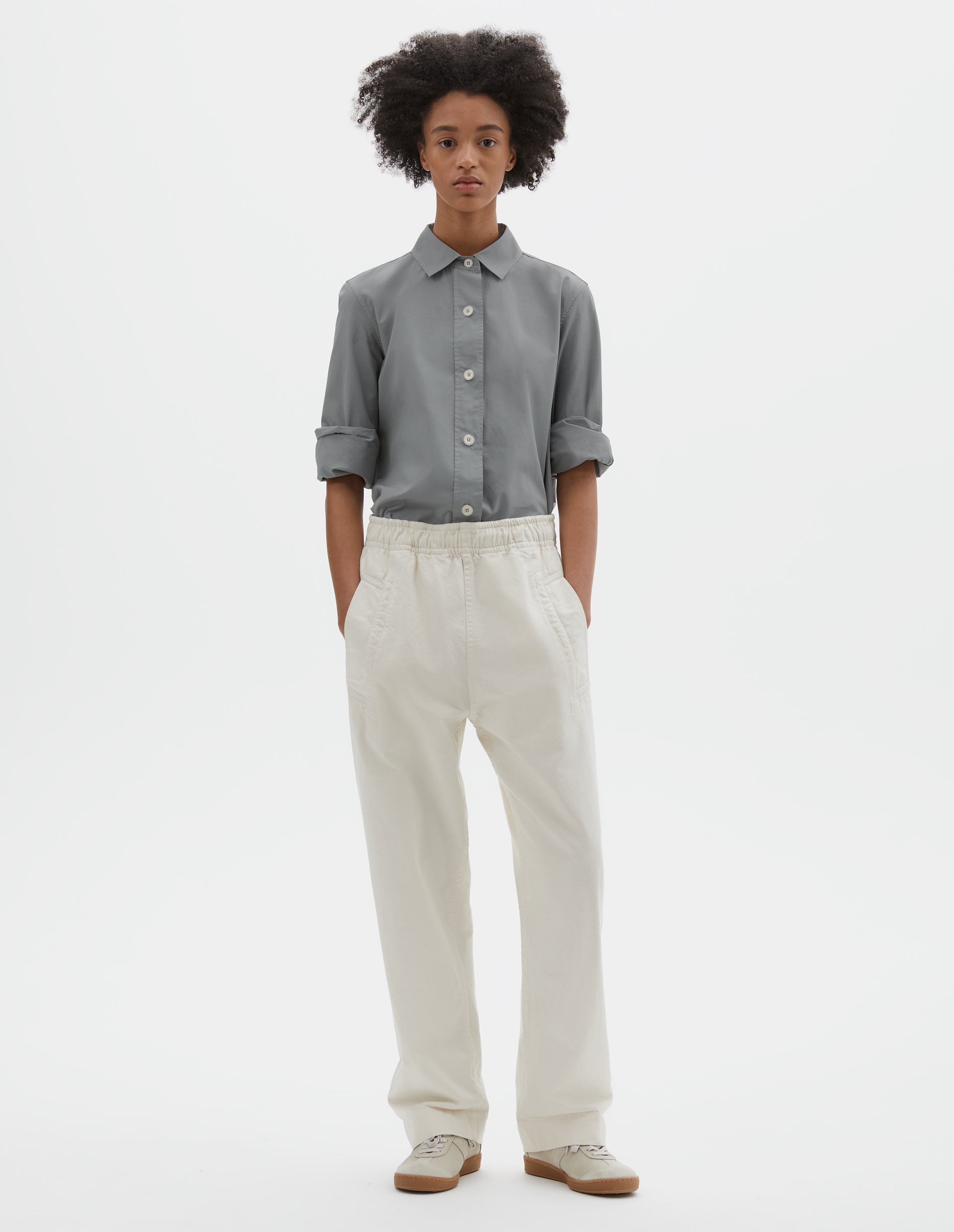 Full body image, front view of model wearing grey regular cut shirt with stitch through raised placket with corozo buttons on placket and cuffs. Styled with beige joggers and beige nubuck trainers.