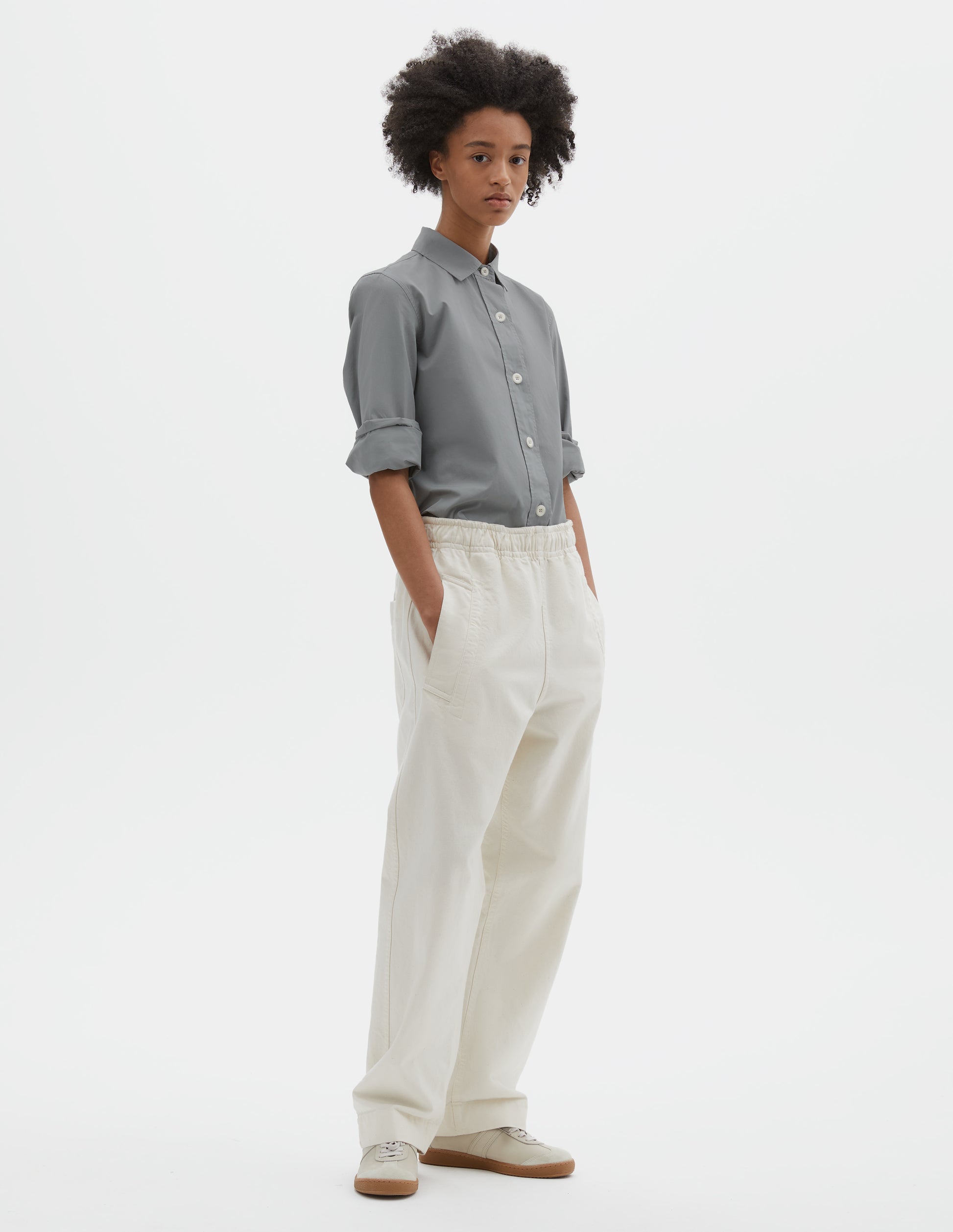 Full body image, front view of model wearing relaxed white joggers with elasticated waist and deep welt pockets. Styled with grey cotton shirt tucked in and off white suede trainers.