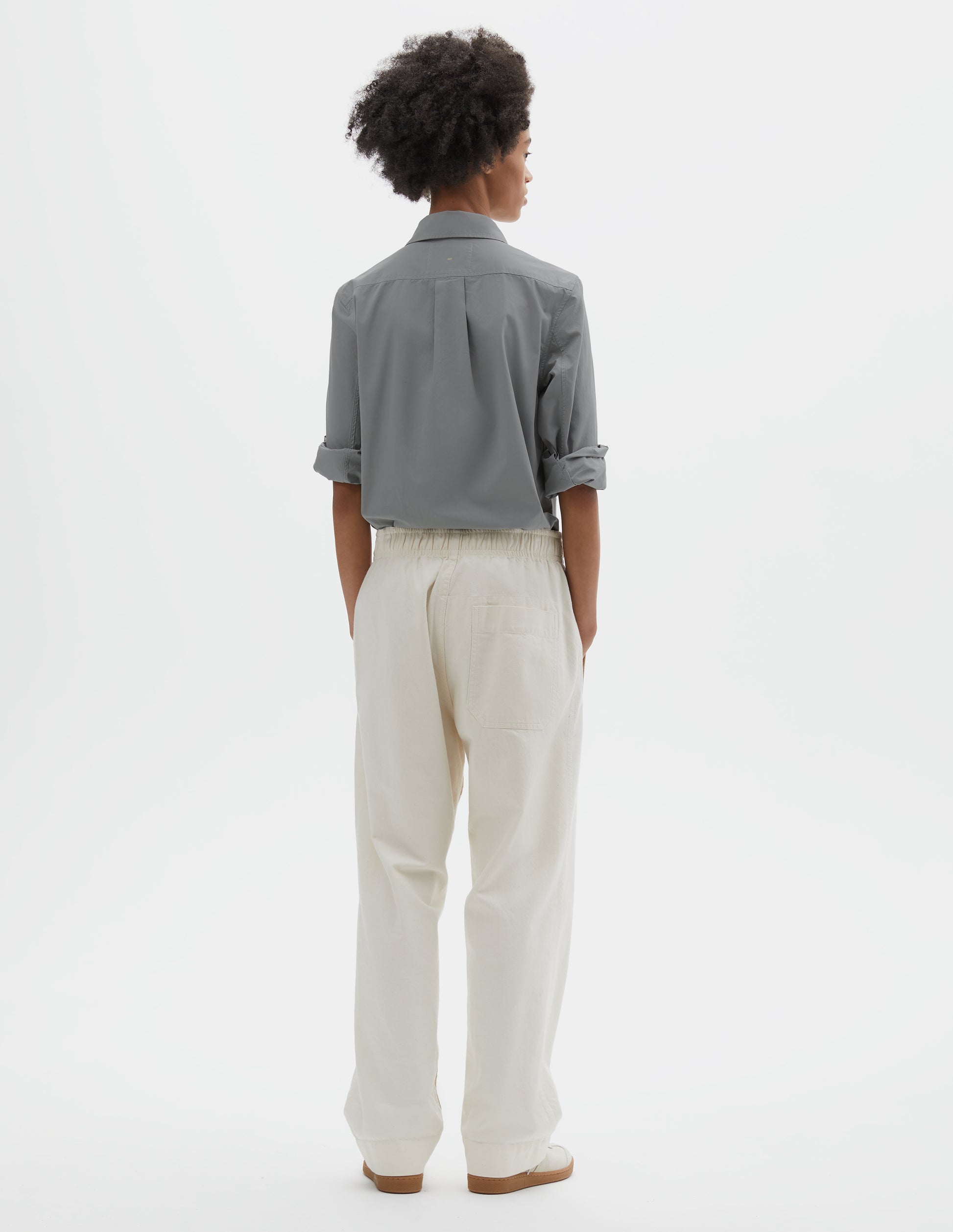Full body image, back view of model wearing relaxed white joggers with elasticated waist and patch back pocket. Styled with grey cotton shirt tucked in and off white suede trainers.
