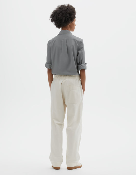 Full body image, back view of model wearing relaxed white joggers with elasticated waist and patch back pocket. Styled with grey cotton shirt tucked in and off white suede trainers.