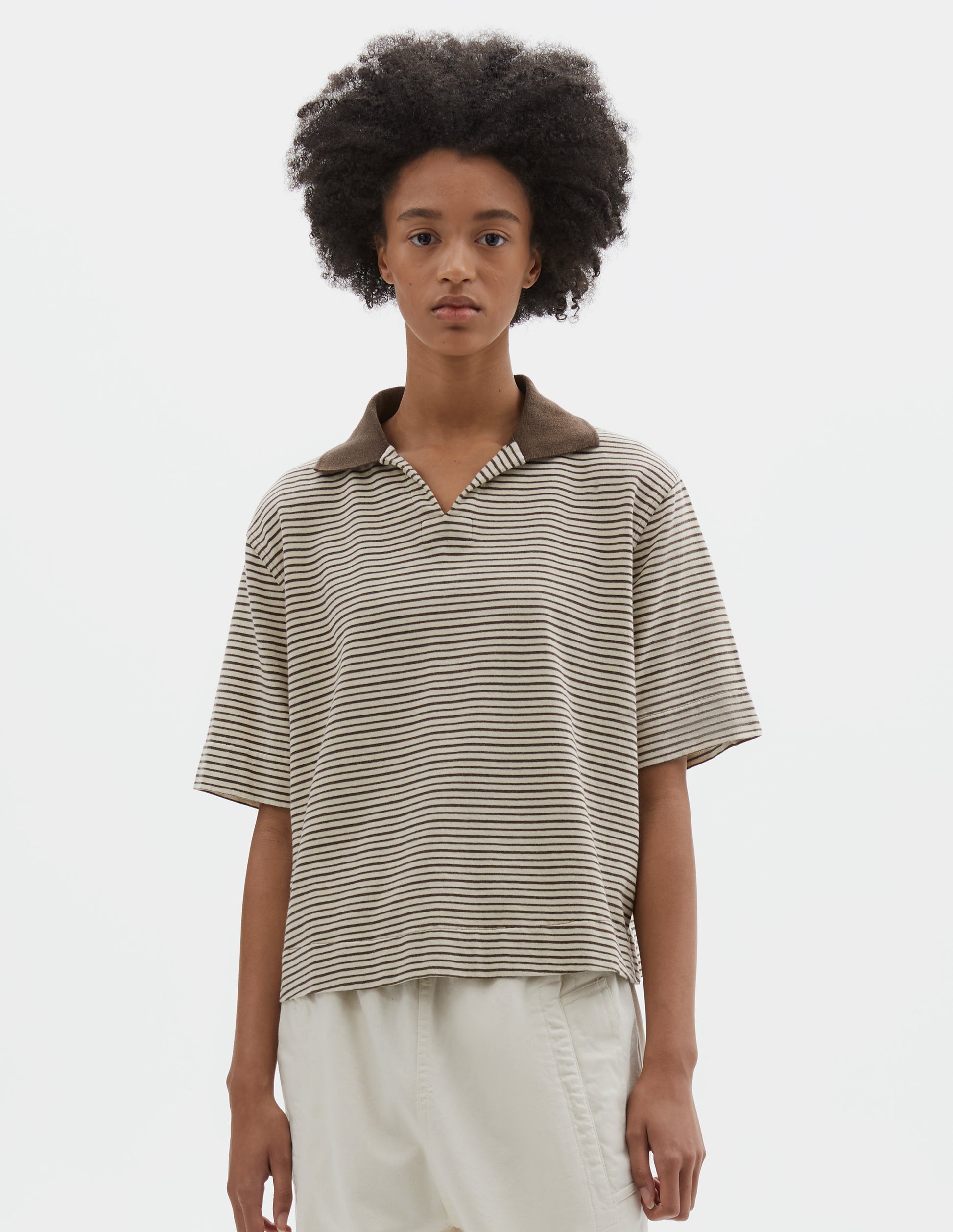 Cropped body image, front view of model wearing brown and white striped short sleeve polo top, with contrast brown ribbed open collar. Styled with off white trousers.