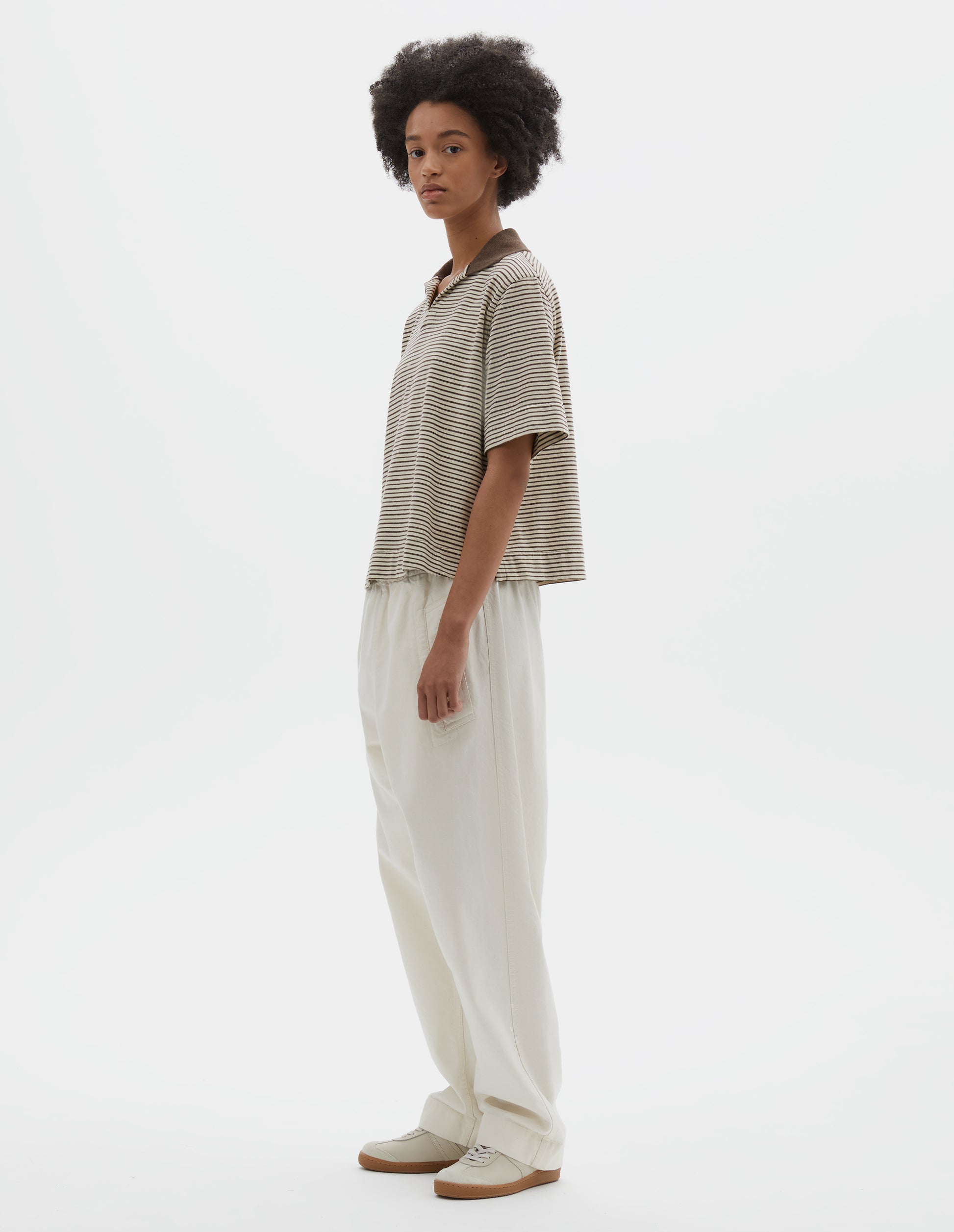 Full body image, side view of model wearing brown and white striped short sleeve polo top, with contrast brown ribbed open collar. Styled with off white trousers and beige suede trainers. 