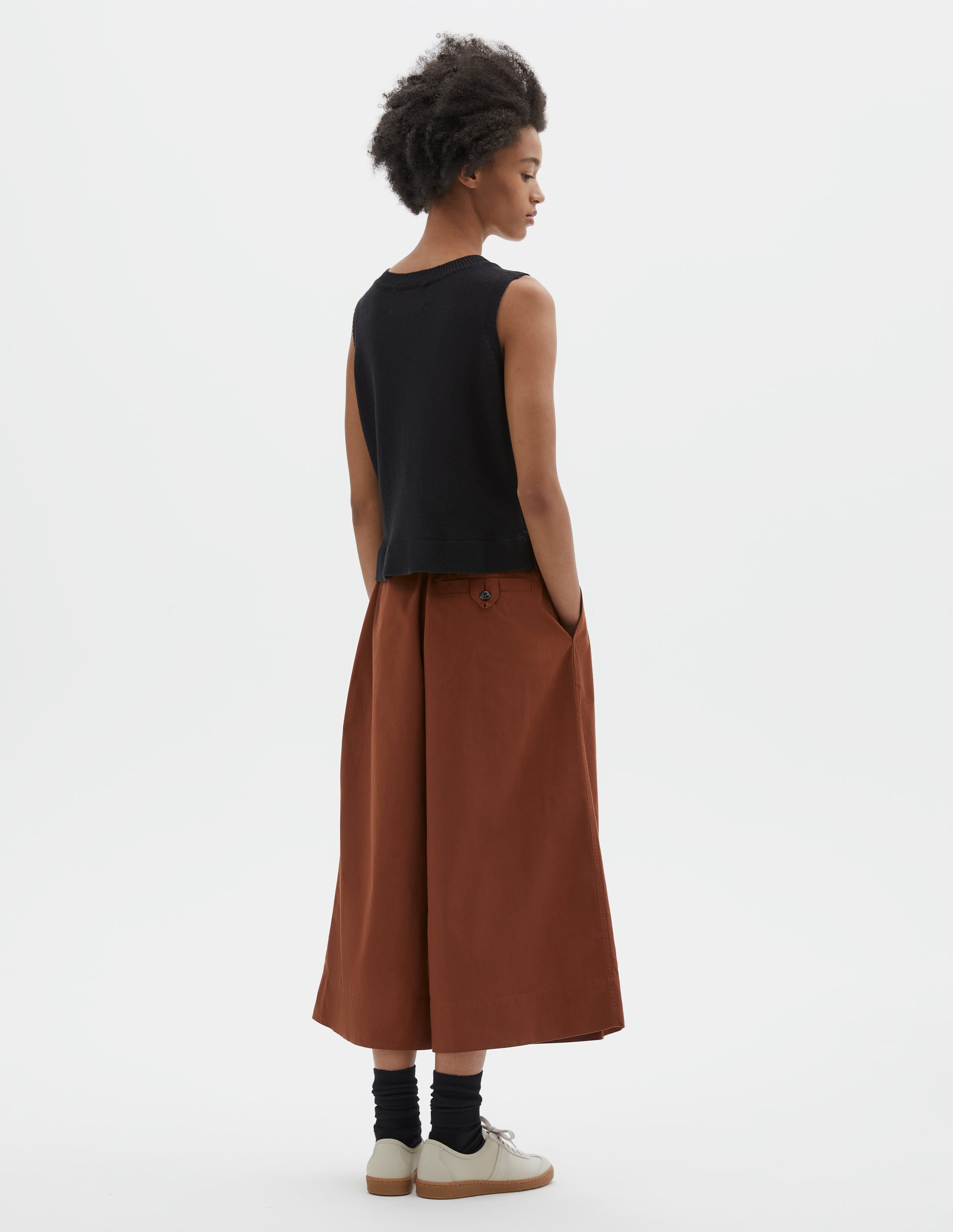 Full body image, back view of model wearing regular fit black knitted tank top with single rib crew neck and side split rib hem. Styled with dark orange culottes and black socks with beige nubuck trainers.