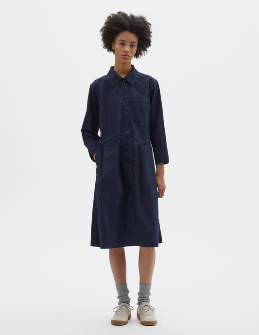 Full body image, front view of model wearing dark blue cotton pleated shirt dress with patch pocket on chest, corozo button fastening, pleats on seam from waist down and side pockets. Styled with light green socks and beige nubuck trainers.