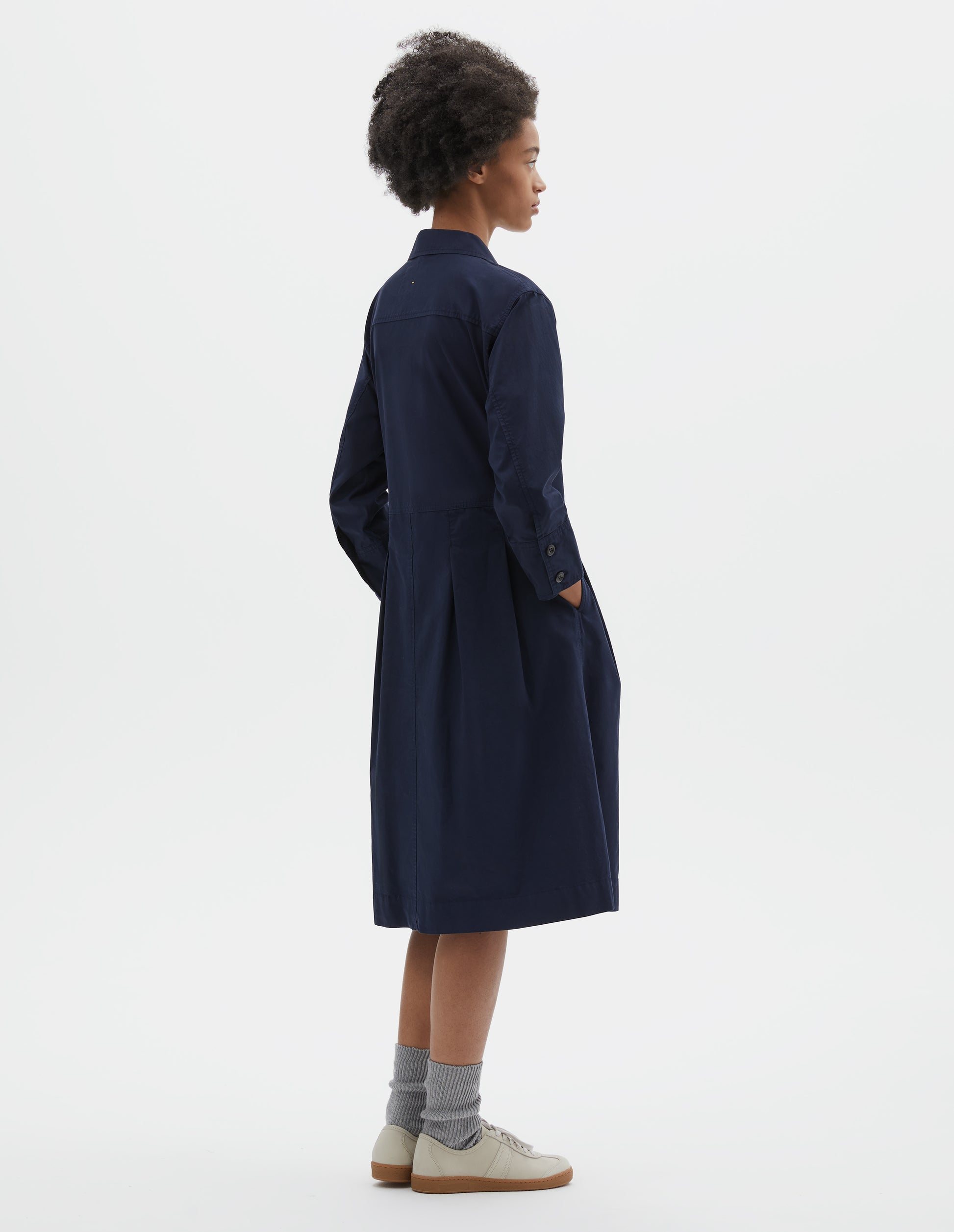 Full body image, back view of model wearing dark blue cotton pleated shirt dress with double corozo button fastening cuffs, pleats on seam from waist down and side pockets. Styled with light green socks and beige nubuck trainers.