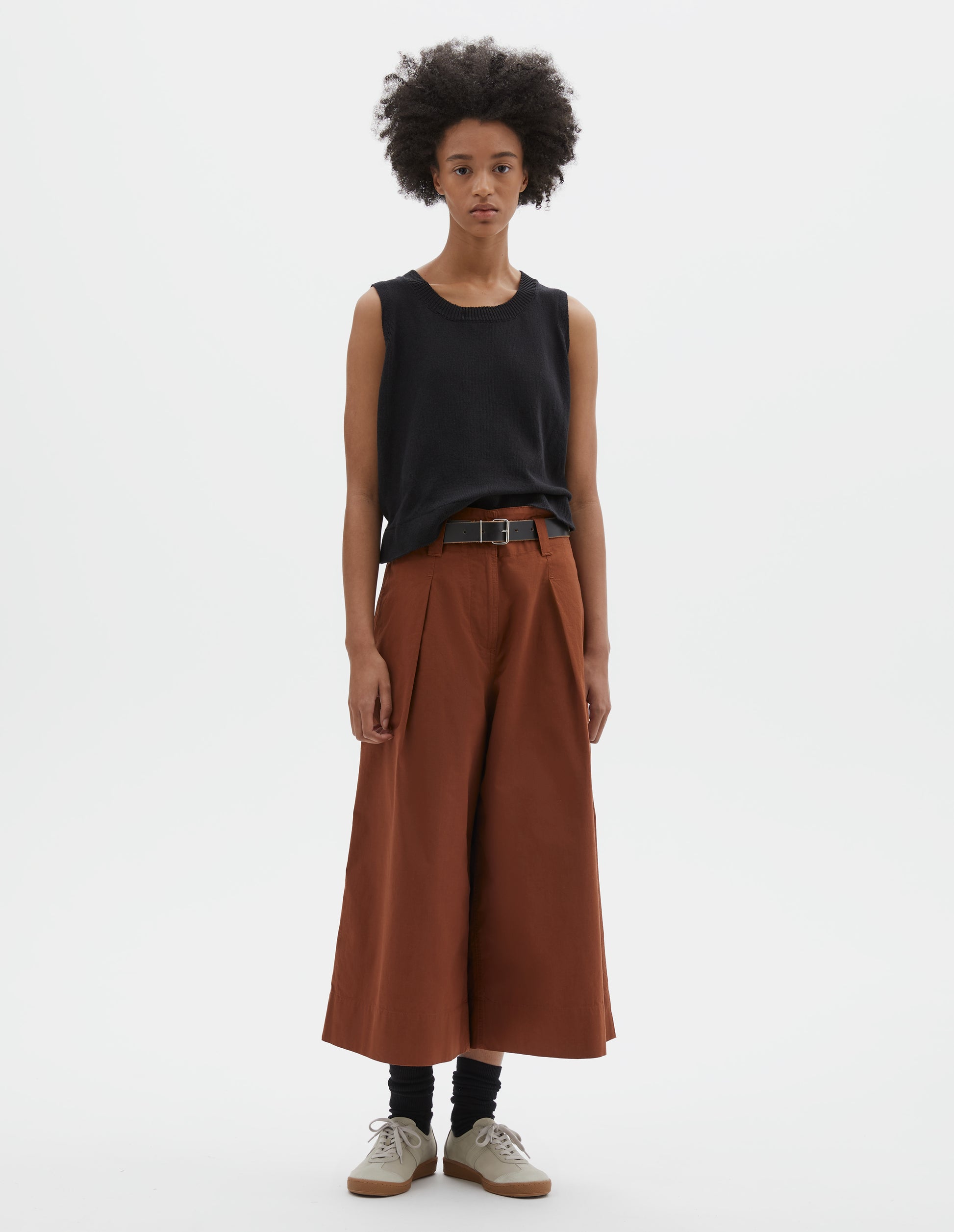 Full body image, front view of model wearing relaxed wide leg dark orange culottes with zip fly, stitch-through waistband with dropped belt loops, bar tacked pleats, concealed wide pockets and deep stitch-through hem. Styled with black sleeveless top, black leather belt, black socks and beige nubuck trainers.