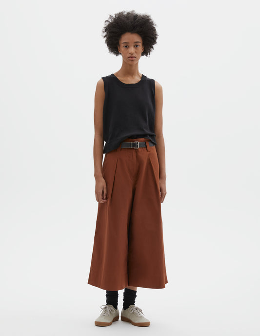 Full body image, front view of model wearing relaxed wide leg dark orange culottes with zip fly, stitch-through waistband with dropped belt loops, bar tacked pleats, concealed wide pockets and deep stitch-through hem. Styled with black sleeveless top, black leather belt, black socks and beige nubuck trainers.