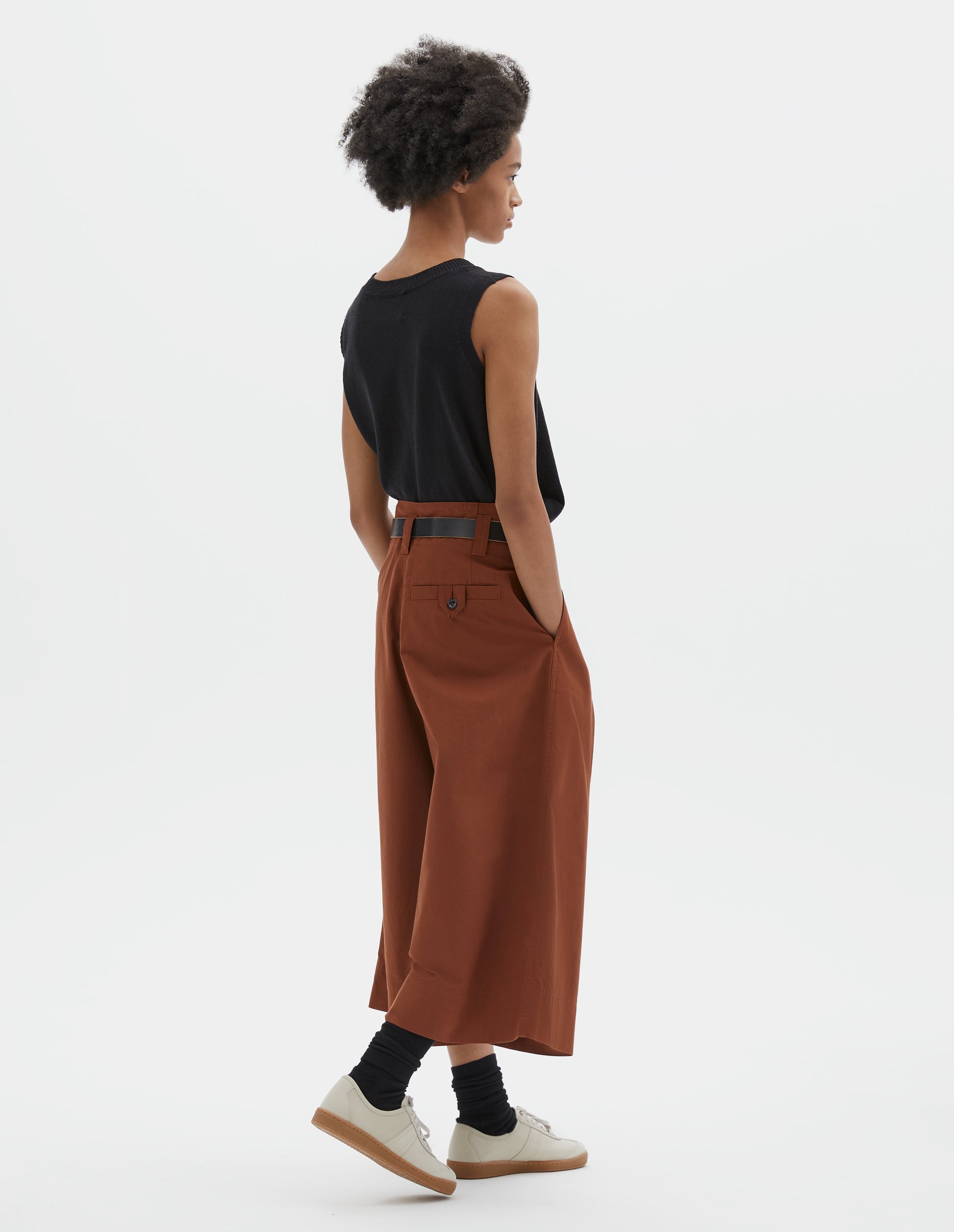 Full body image, back view of model wearing relaxed wide leg dark orange culottes with stitch-through waistband and dropped belt loops, and corozo button loop fastening welt pocket. Styled with black sleeveless top, black leather belt, black socks and beige nubuck trainers.