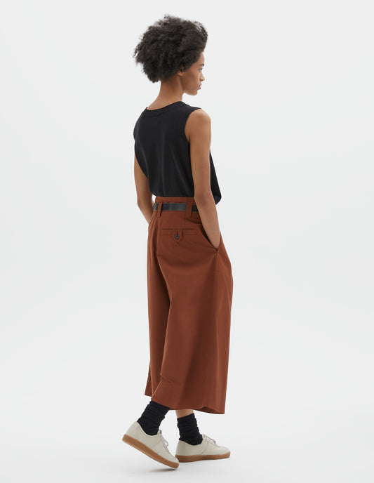 Full body image, back view of model wearing relaxed wide leg dark orange culottes with stitch-through waistband and dropped belt loops, and corozo button loop fastening welt pocket. Styled with black sleeveless top, black leather belt, black socks and beige nubuck trainers.
