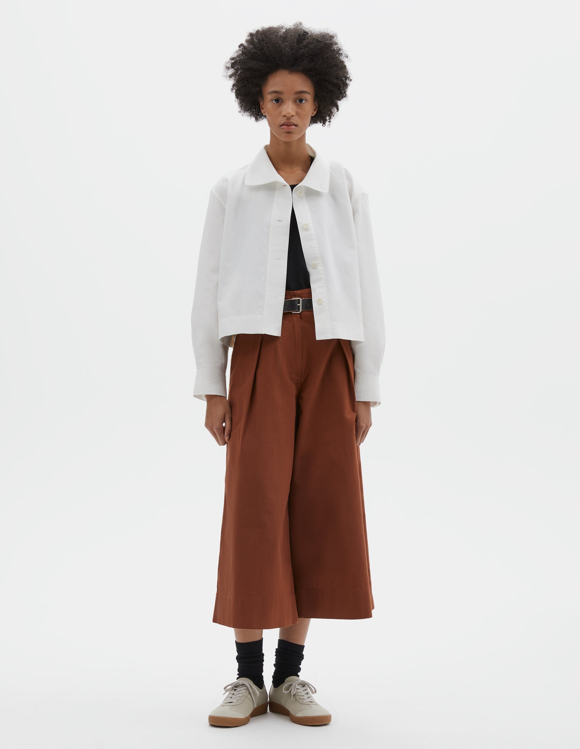 Full body image, front view of model wearing boxy cropped white shirt with round asymmetrical collar, wide stitch-through placket with corozo brace buttons and stepped vent split hem.  Styled over black top and  with black leather belt, dark orange culottes and  black socks with beige nubuck trainers.