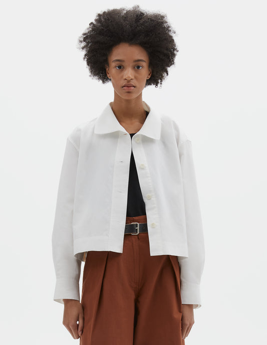 Cropped body image, front view of model wearing boxy cropped white shirt with round asymmetrical collar, wide stitch-through placket with corozo brace buttons and stepped vent split hem.  Styled over black top, with black leather belt and dark orange culottes.