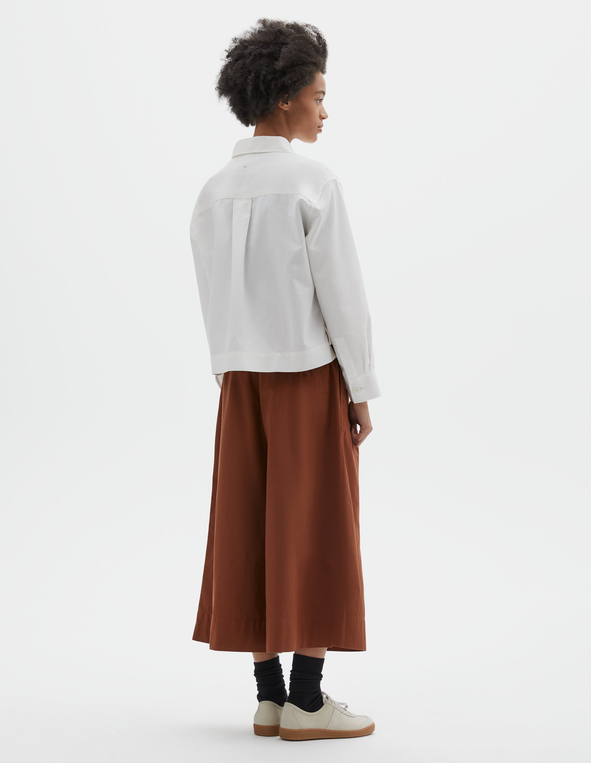 Full body image, back view of model wearing boxy cropped white shirt with stepped vent split hem, corozo brace buttons on cuffs and box pleat on back yoke. Styled with dark orange culottes and black socks with beige nubuck trainers.