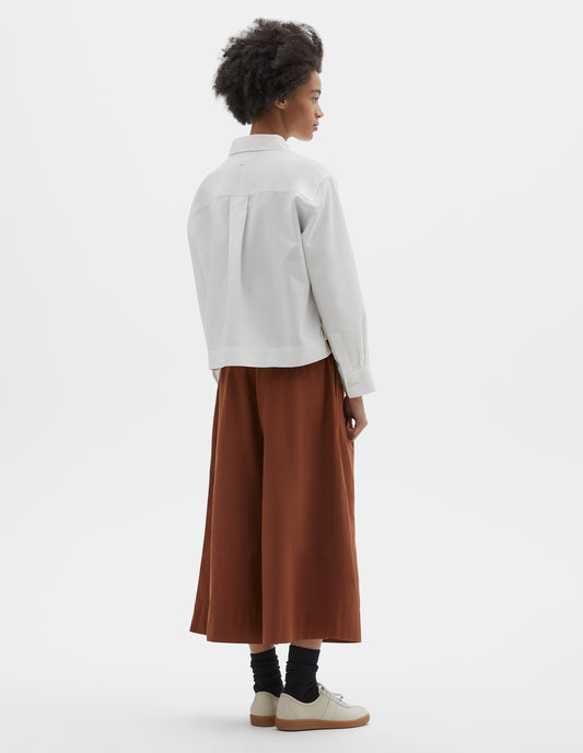 Full body image, back view of model wearing boxy cropped white shirt with stepped vent split hem, corozo brace buttons on cuffs and box pleat on back yoke. Styled with dark orange culottes and black socks with beige nubuck trainers.