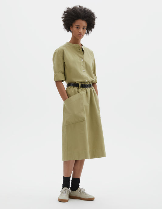 Full body image, front view of model wearing relaxed light green skirt with two large patch pockets, elasticated waist and belt loops. Styled with pale green shirt, black leather belt, black socks and beige nubuck trainers.