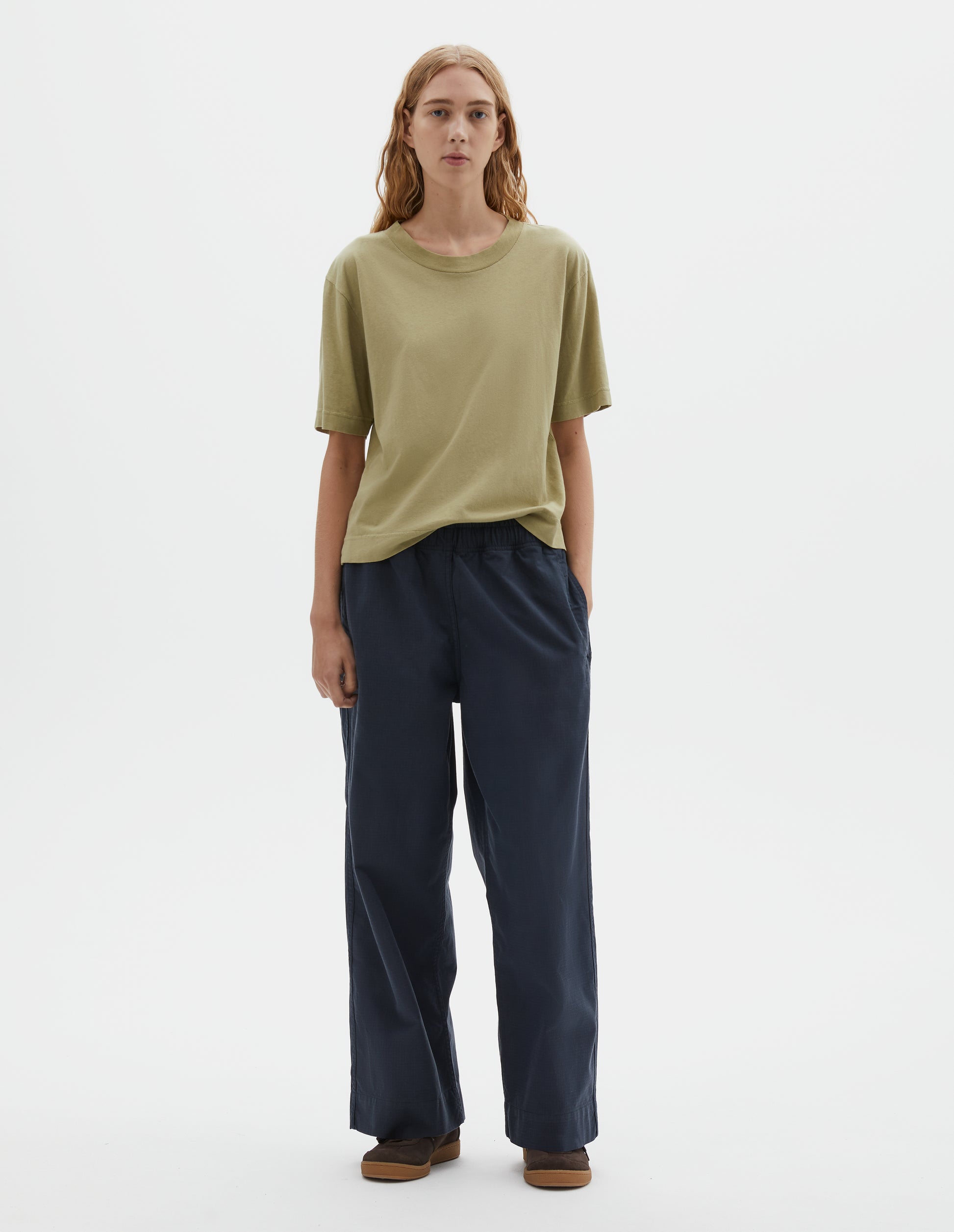 Full body image, front view of model wearing dark navy blue joggers with elasticated waistband and side seam pockets. Styled with pale green cotton t-shirt and brown suede trainers.