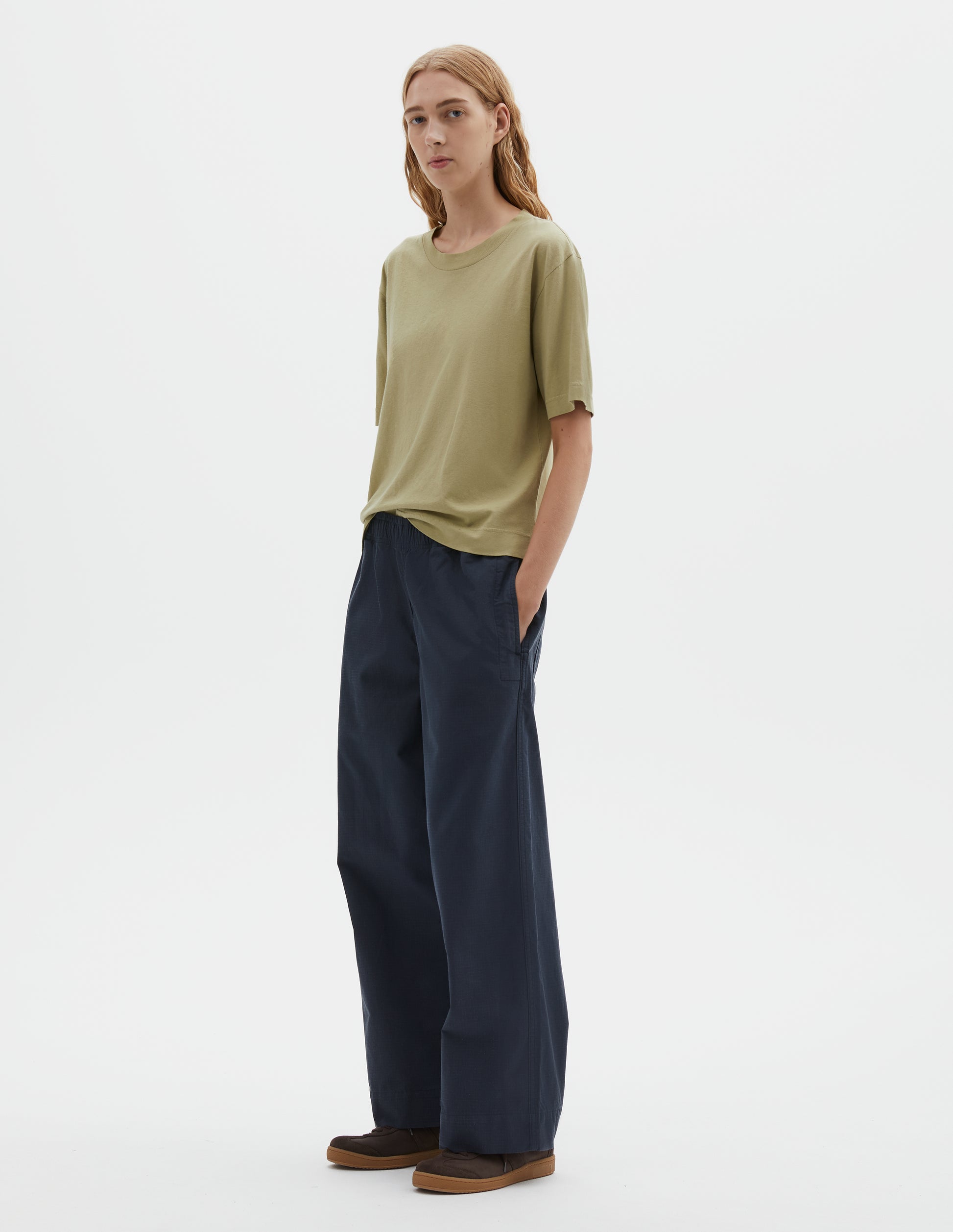 Full body image, front view of model wearing pale green cotton linen short sleeve t-shirt with vent split hem. Styled with dark navy wide leg trouser and brown suede trainers.