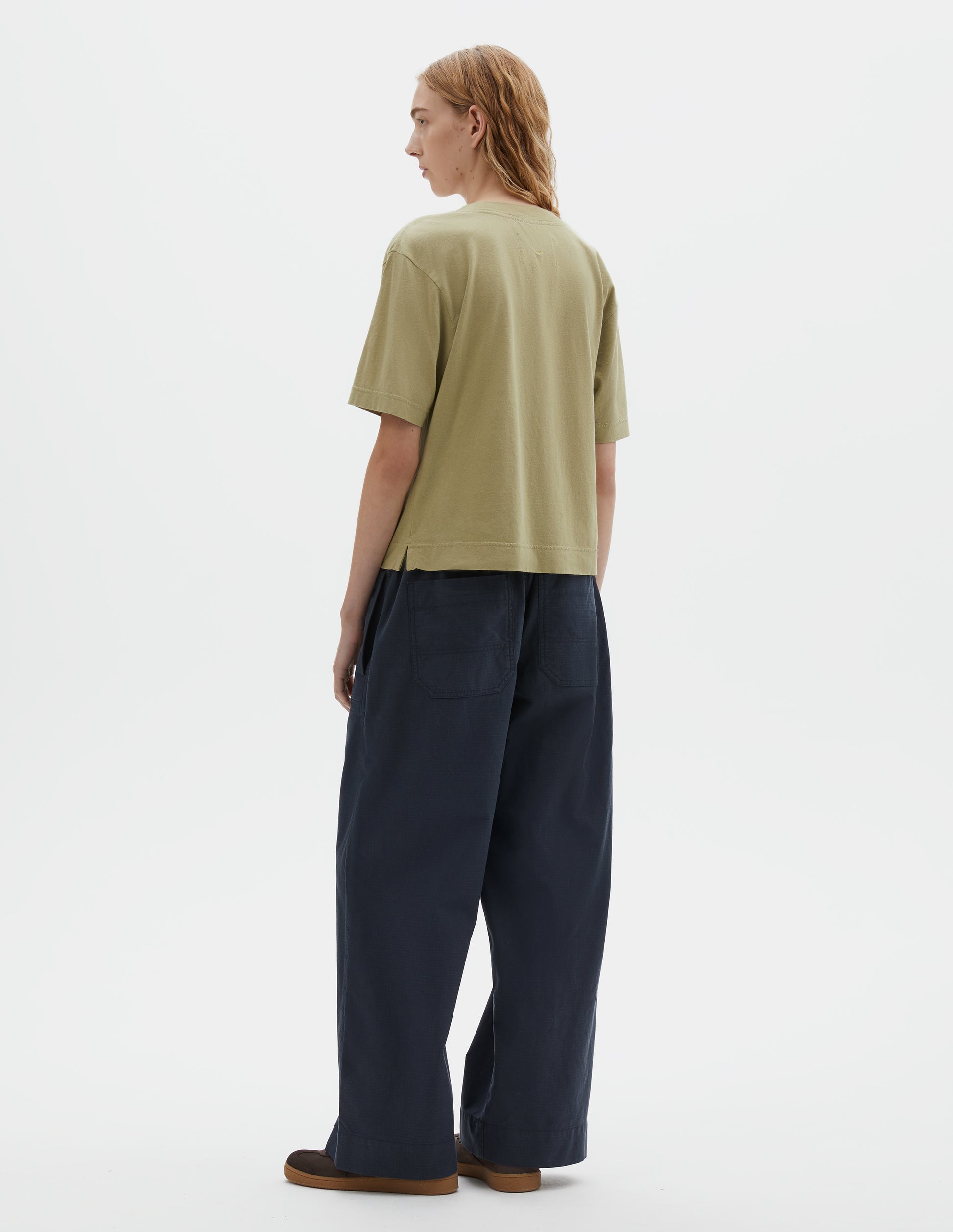 Full body image, back view of model wearing pale green cotton linen short sleeve t-shirt with vent split hem. Styled with dark navy wide leg trouser and brown suede trainers.
