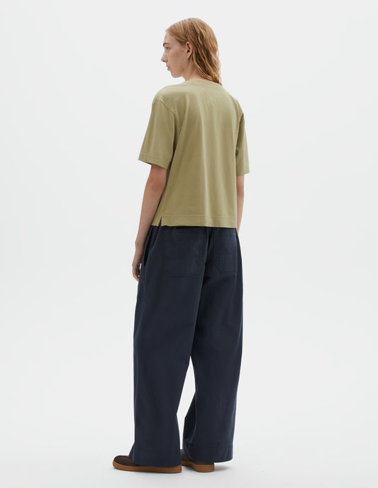 Full body image, back view of model wearing pale green cotton linen short sleeve t-shirt with vent split hem. Styled with dark navy wide leg trouser and brown suede trainers.