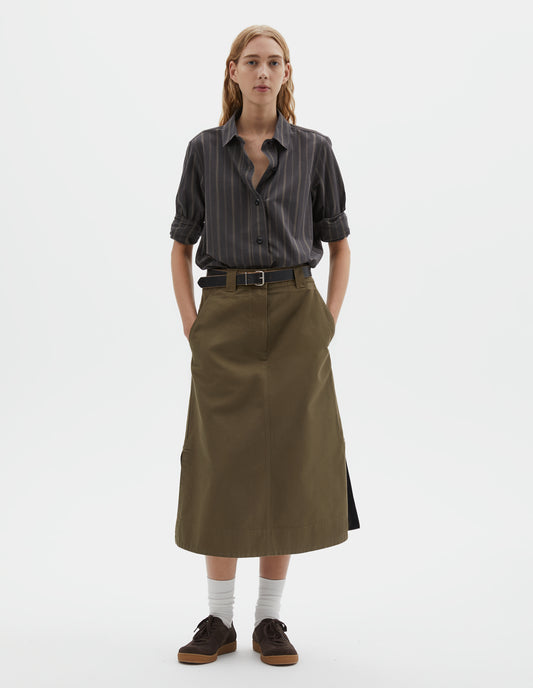 Full body image, front view of model wearing off black and khaki striped shirt, with corozo button fastening. Styled tucked in to green cotton skirt, with black leather belt, white socks and brown suede trainers. 