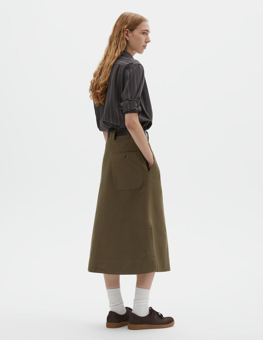 Full body image, back view of model wearing green regular cut long length A line skirt with side splits, belt loop detail on back and back patch pocket with reinforced stitch detailing. Styled with black striped shirt, black leather belt, white socks and dark brown nubuck trainers. 