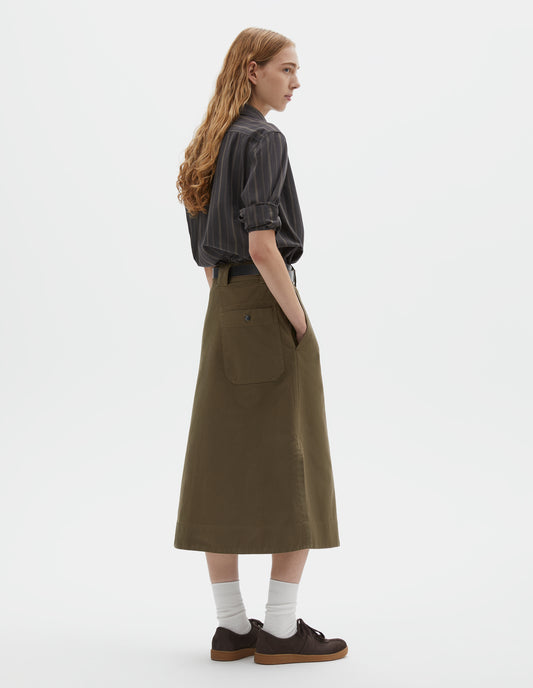 Full body image, back view of model wearing off black and khaki striped shirt with box pleat on back yoke. Styled tucked in to green cotton skirt, with black leather belt, white socks and brown suede trainers. 