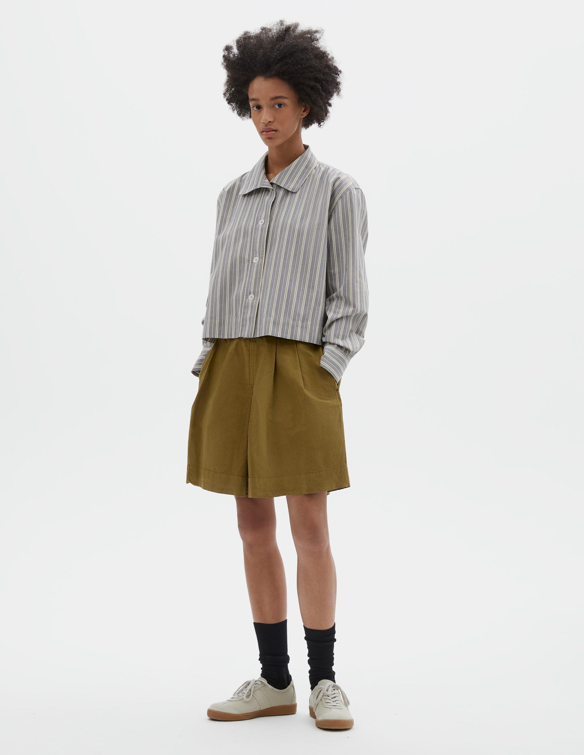 Full body image, front view of model wearing grey, yellow and white striped cotton long sleeve shirt. With round collar, wide stitch through placket, stepped vent split hem and corozo brace button closure. Styled with green pleated shorts, black socks and beige suede trainers.
