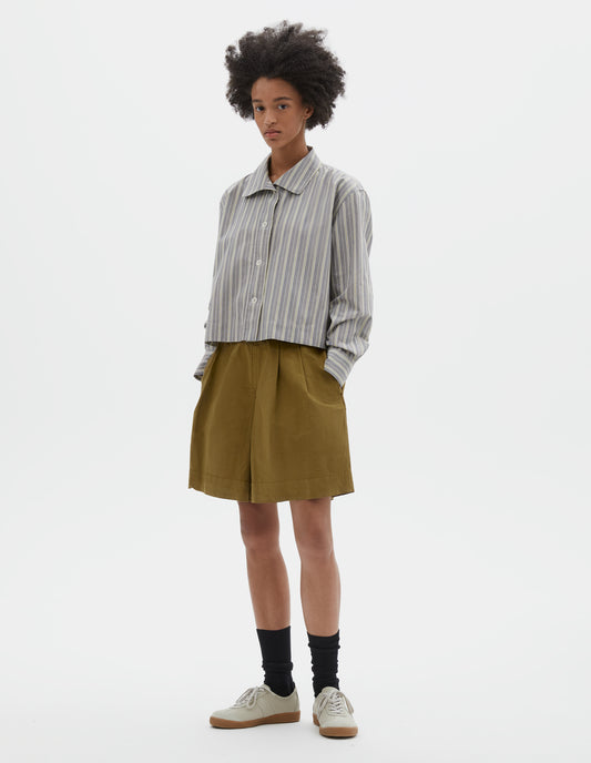 Full body image, front view of model wearing grey, yellow and white striped cotton long sleeve shirt. With round collar, wide stitch through placket, stepped vent split hem and corozo brace button closure. Styled with green pleated shorts, black socks and beige suede trainers.