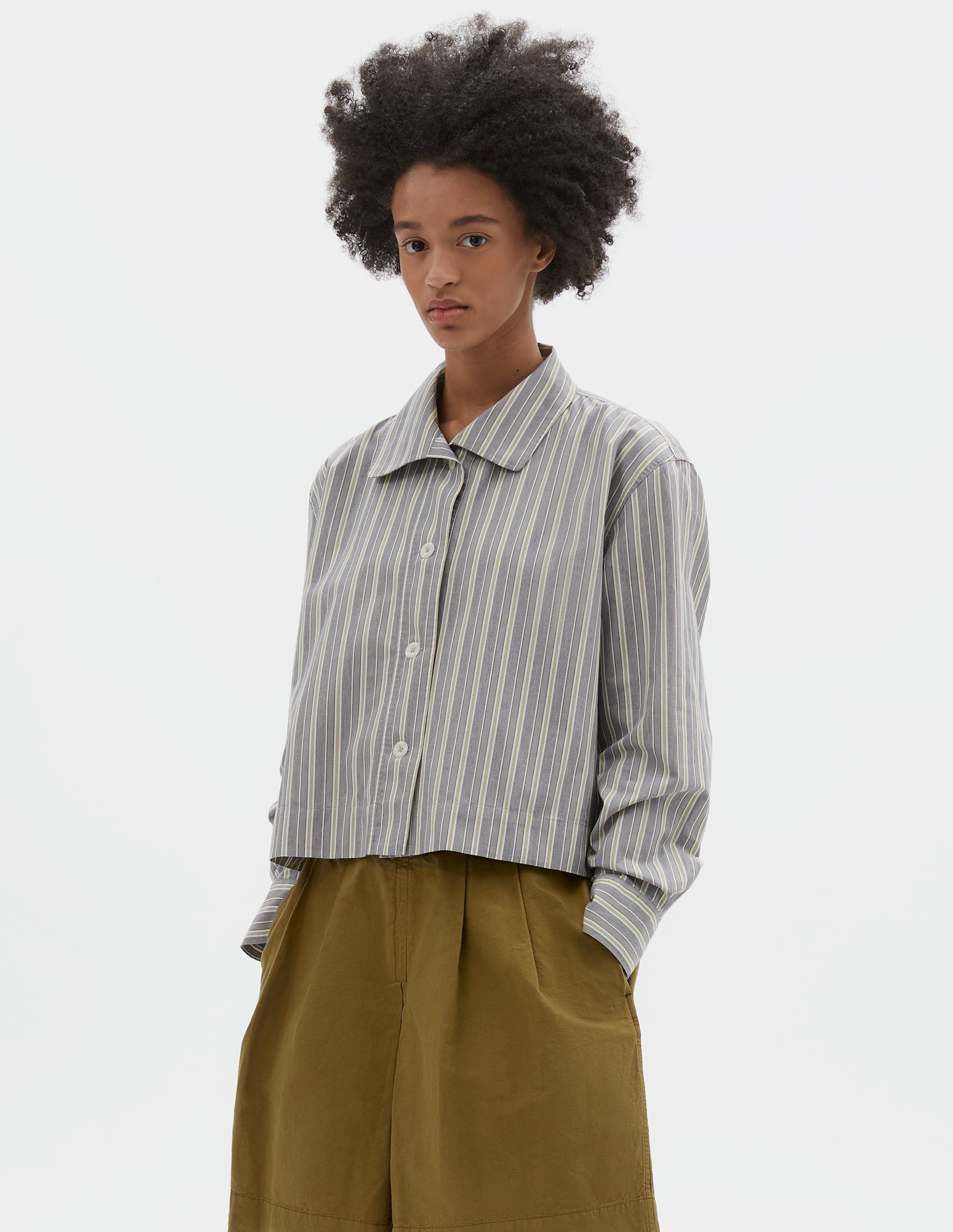 Cropped body image, front view of model wearing grey, yellow and white striped cotton long sleeve shirt. With round collar, wide stitch through placket, stepped vent split hem and corozo brace button closure. Styled with green pleated shorts.
