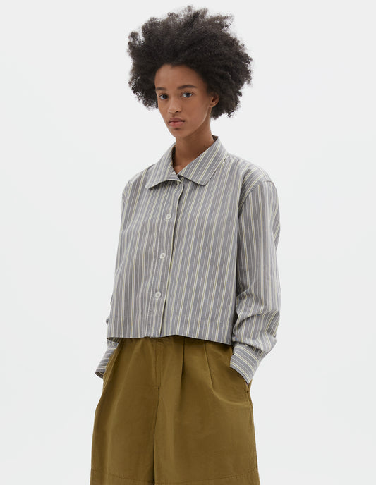 Cropped body image, front view of model wearing grey, yellow and white striped cotton long sleeve shirt. With round collar, wide stitch through placket, stepped vent split hem and corozo brace button closure. Styled with green pleated shorts.